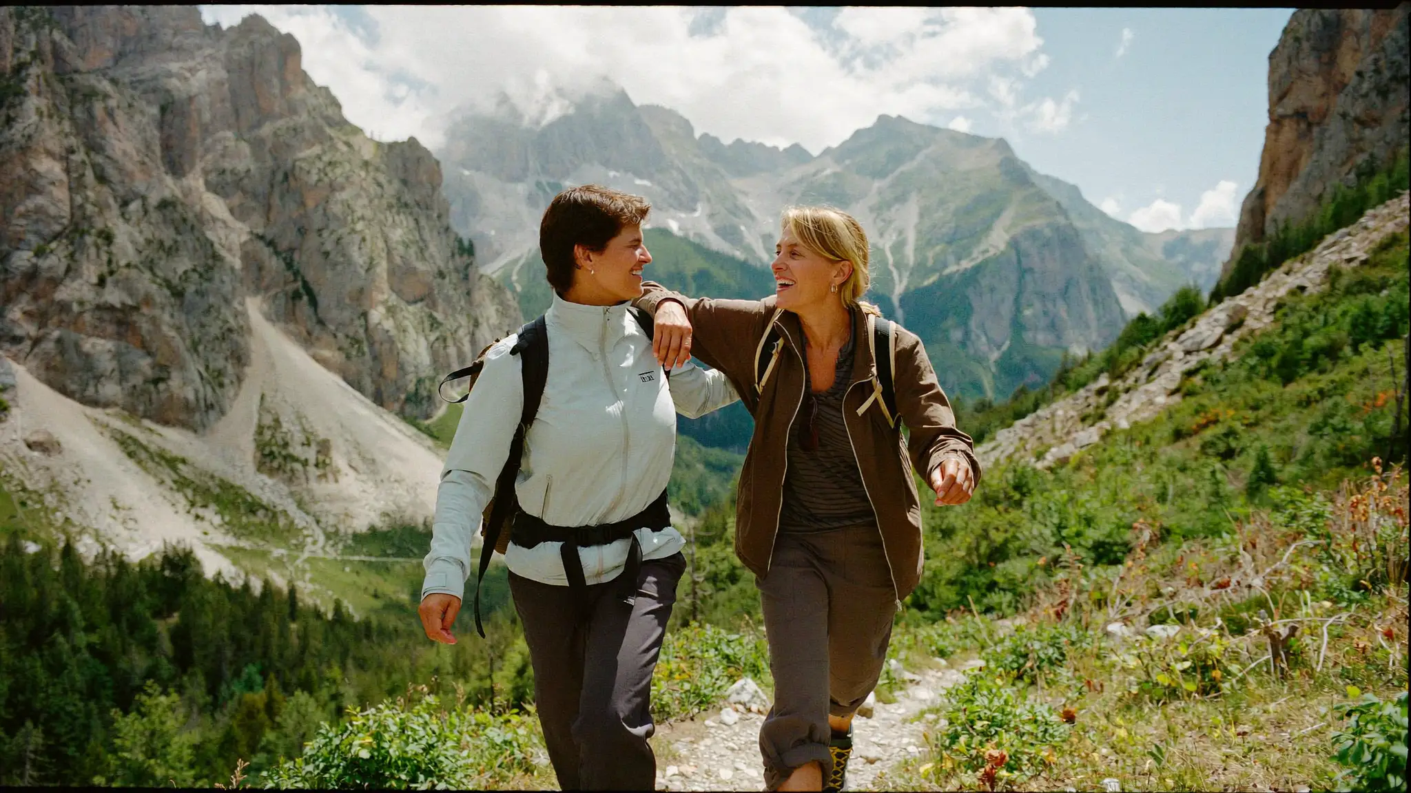 Zwei glückliche Frauen beim gemeinsamen Wandern auf einem Bergweg in den Alpen, einander anlächelnd vor einem Bergpanorama